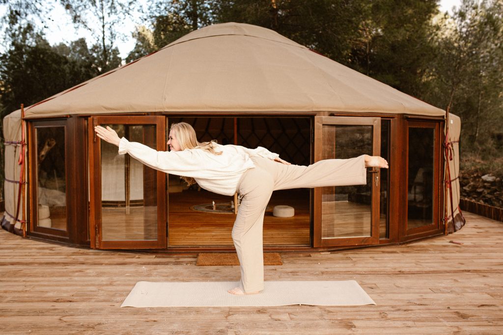 A woman performing a yoga pose on a mat in front of a yurt, surrounded by trees. She is balancing on one leg with her arms extended.