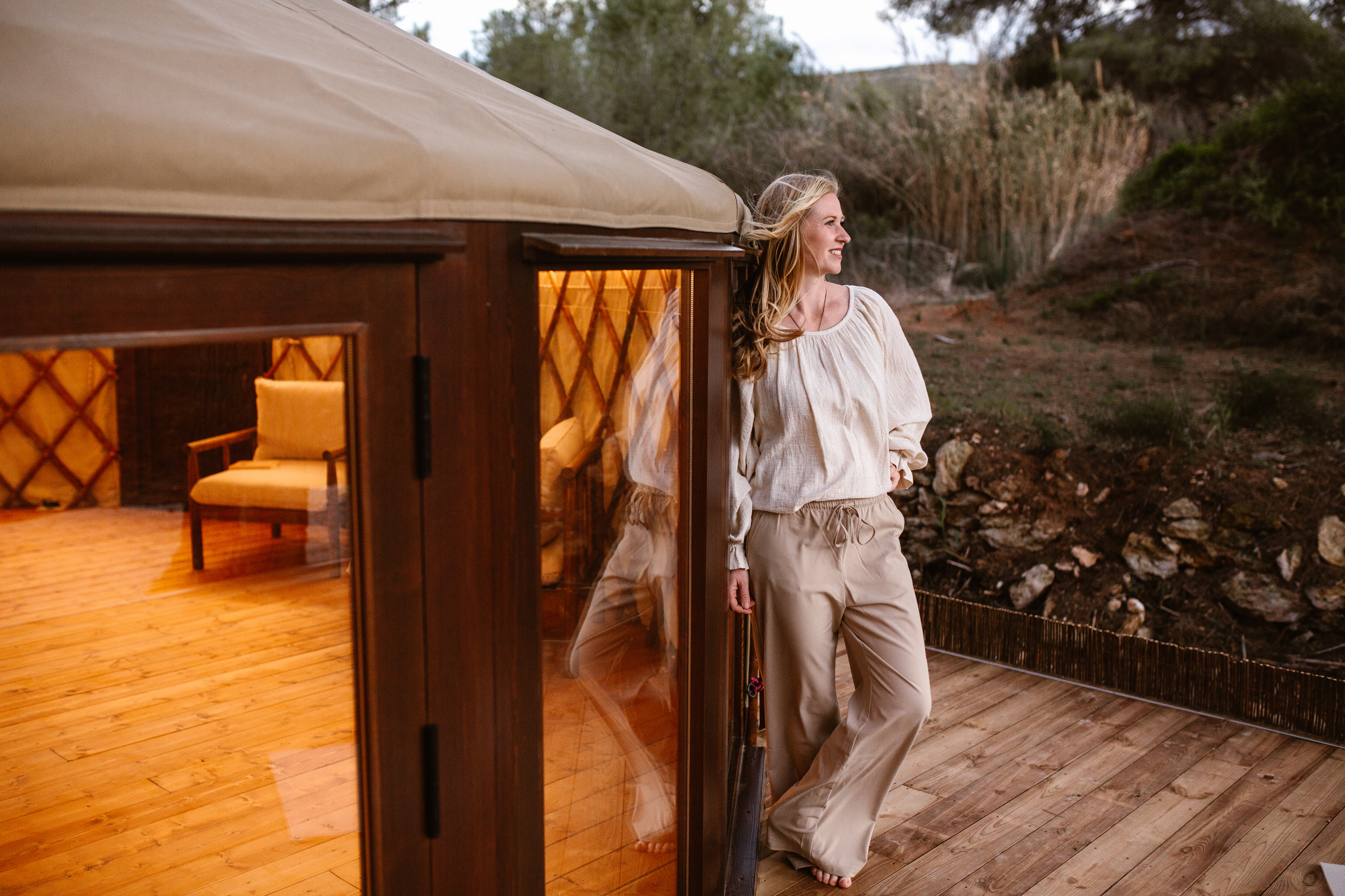 A woman standing beside a wooden structure with warm indoor lighting, wearing a light-colored top and beige pants, smiling and looking off into the distance.