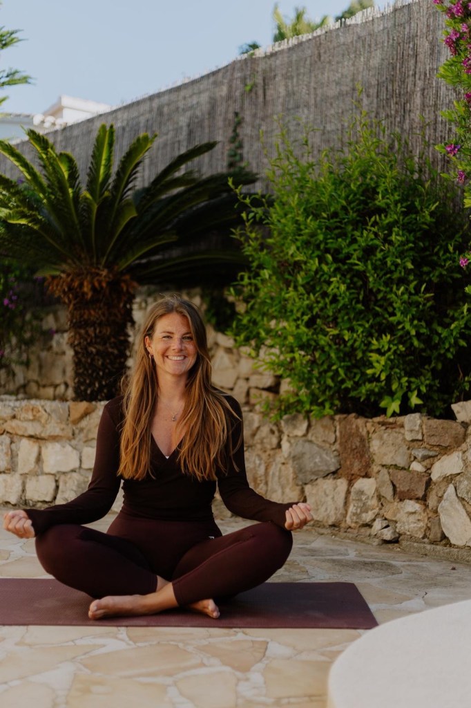 A woman sitting cross-legged on a yoga mat outdoors, smiling and practicing mindfulness in a garden with lush greenery and stone walls.