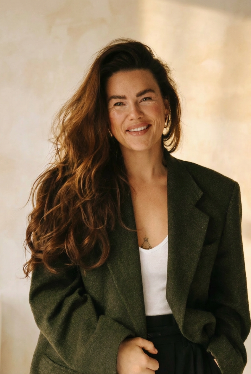 8991D642-B817-4F81-B124-7FF59AE5B9C7 A smiling woman with long, wavy brown hair wearing a green oversized blazer and a white top. She is posing in a softly lit environment with a light beige background.