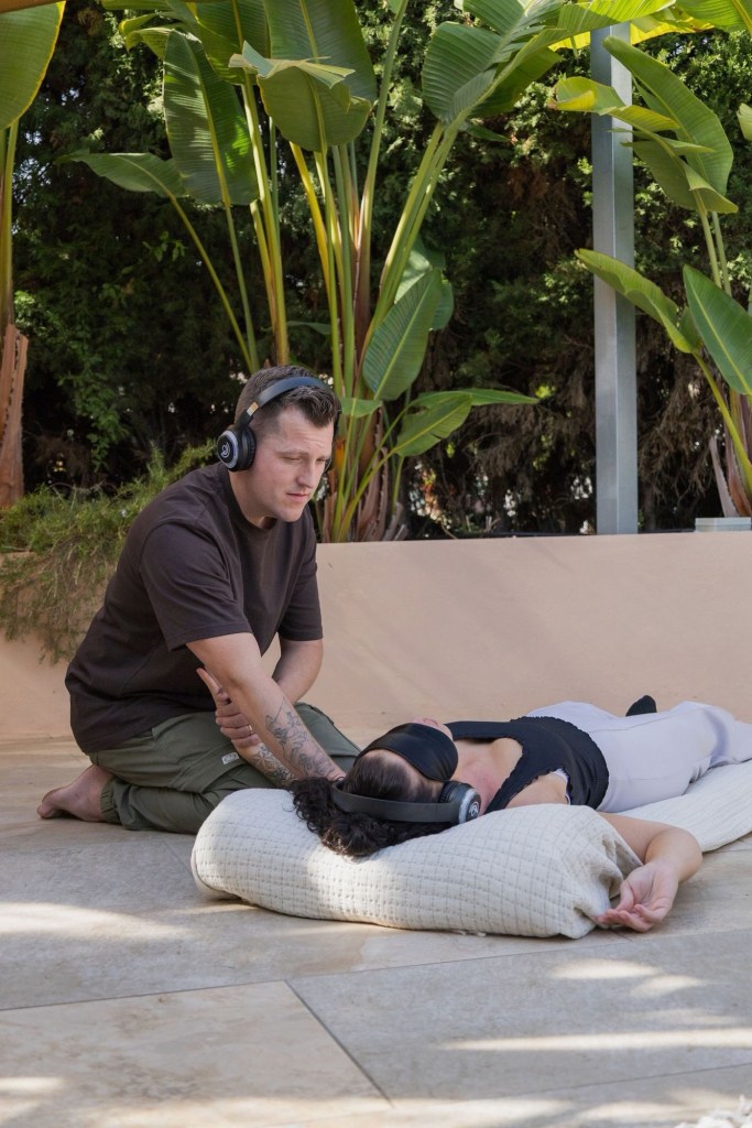 A person sitting on the ground wearing headphones, providing support or guidance to another person lying down with a blindfold and headphones, in a serene outdoor setting surrounded by large green plants.